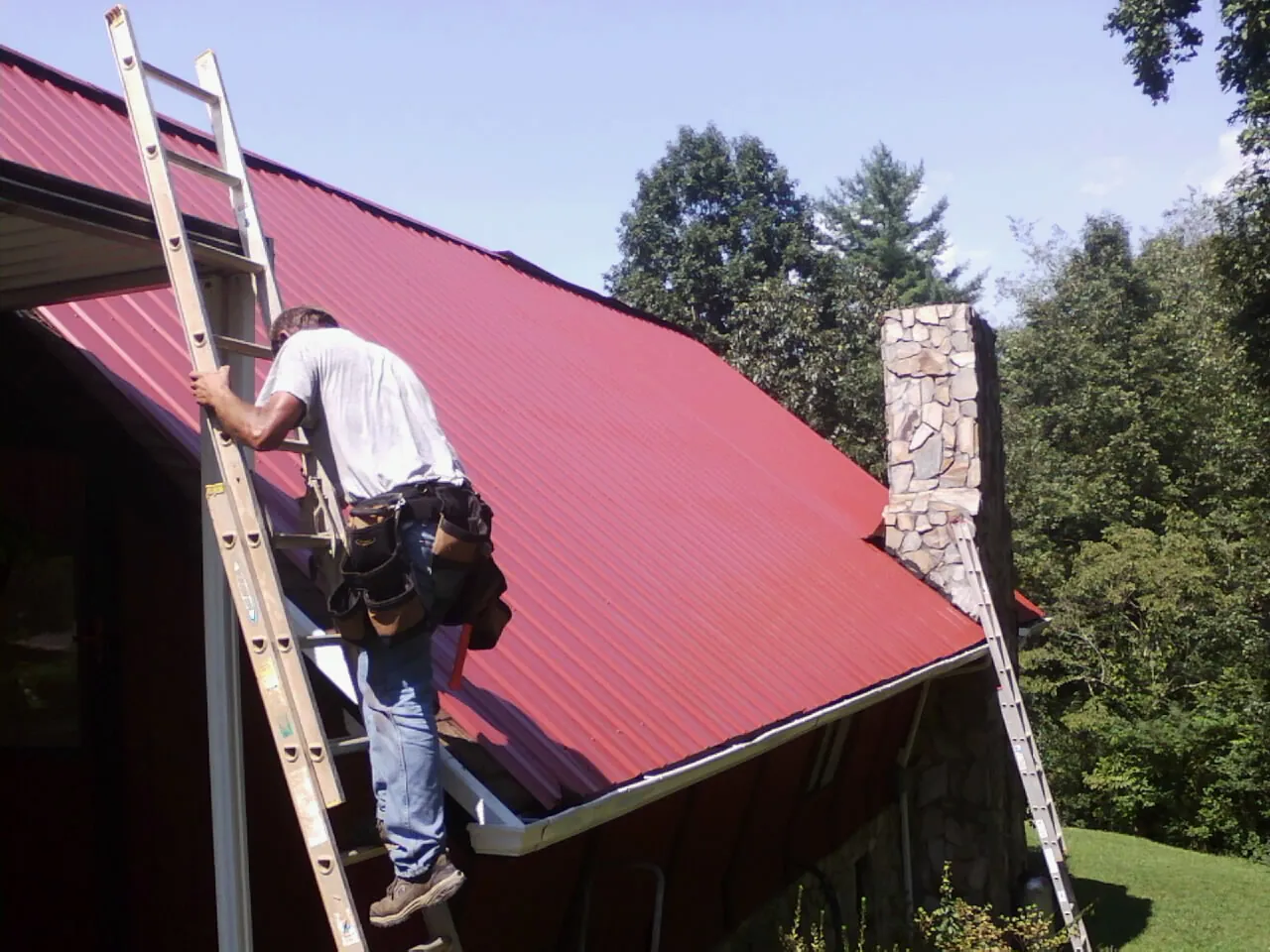 Roofer on a ladder beside a red metal roof and stone chimney — Soffit & Fascia Repair by Roofing Brothers in Beach Haven